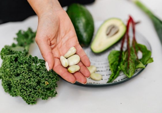 Close-up of a hand holding garlic cloves over a plate with fresh vegetables, including kale and avocado.