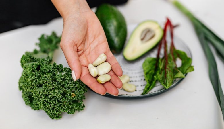 Close-up of a hand holding garlic cloves over a plate with fresh vegetables, including kale and avocado.