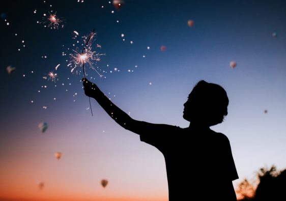 Silhouette of a person holding sparklers against a colorful twilight sky.