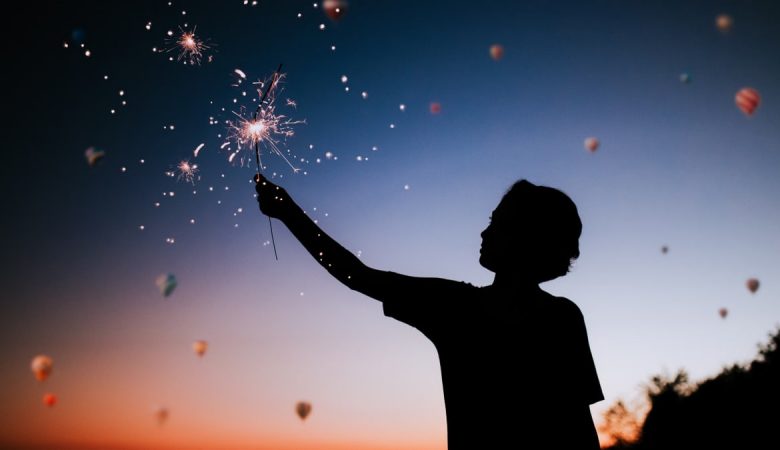 Silhouette of a person holding sparklers against a colorful twilight sky.
